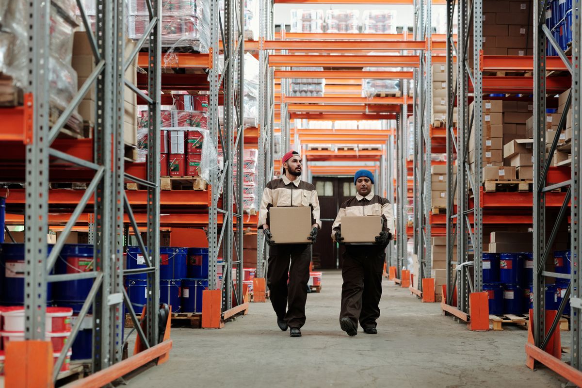 Zwei Mitarbeiter tragen Pakete durch einen Regalgang im Fulfillment-Center